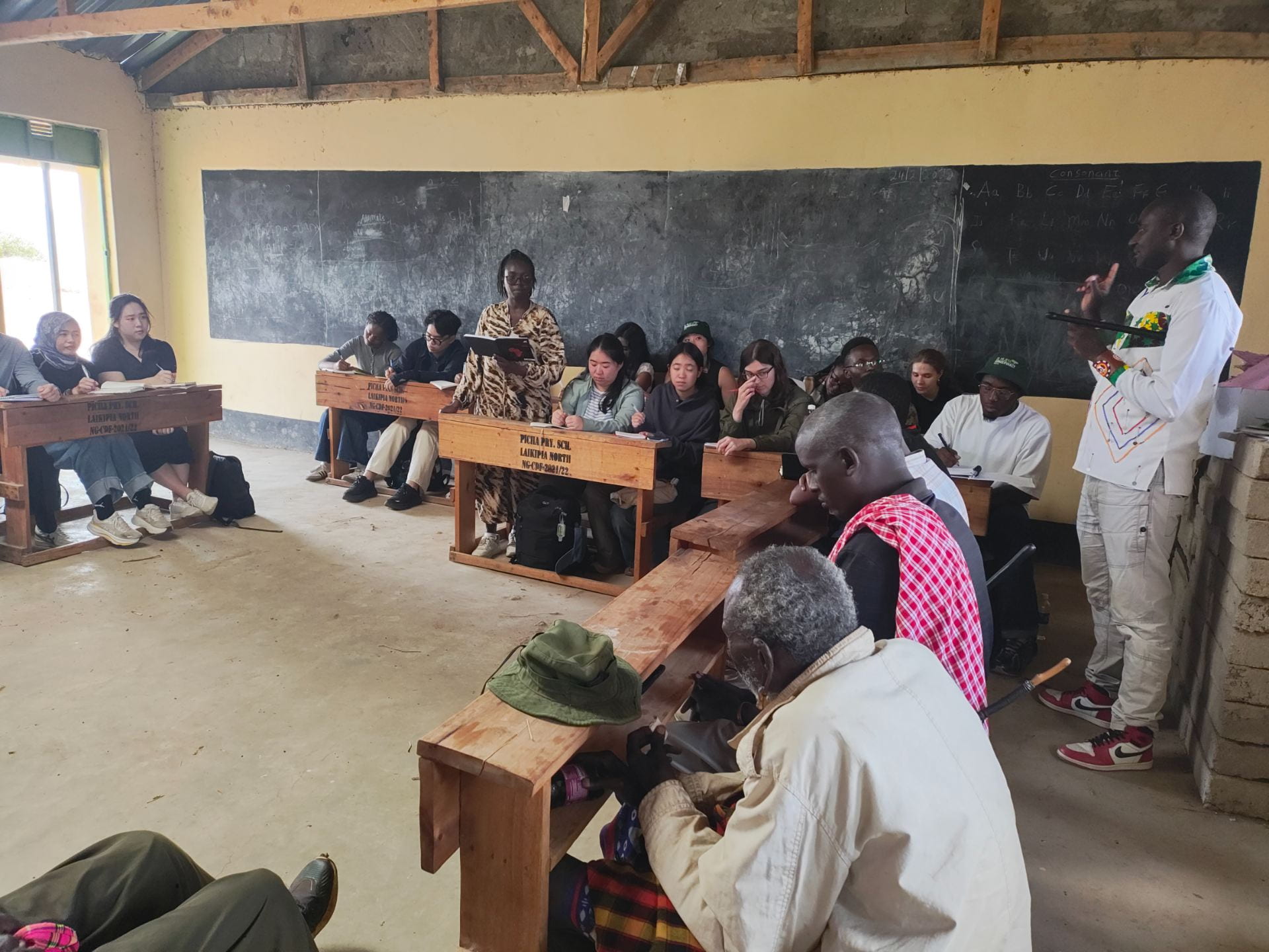 Community meeting in a Laikipia North classroom, with fellows and community members seated at wooden desks